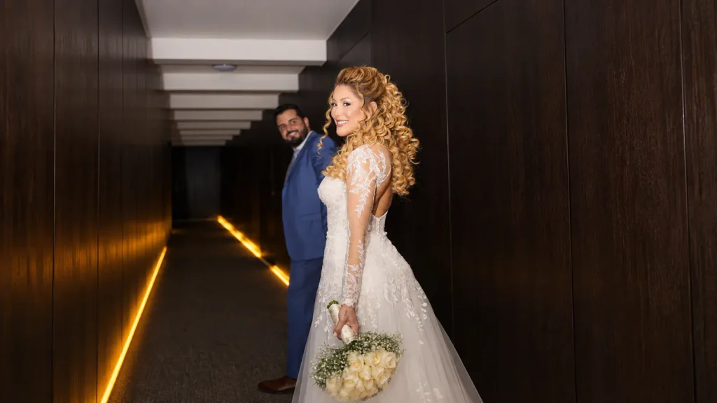 Noiva sorrindo com vestido rendado segurando buquê branco e olhando para trás de mãos dadas com o noivo em terno azul.