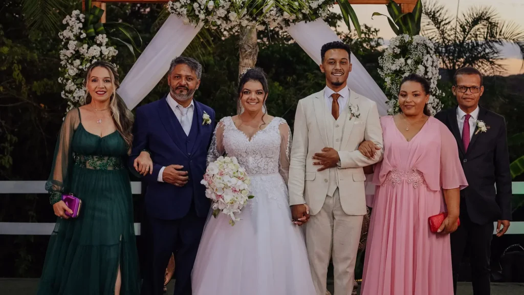Noivos posando ao lado dos pais sob um altar decorado com flores durante o casamento